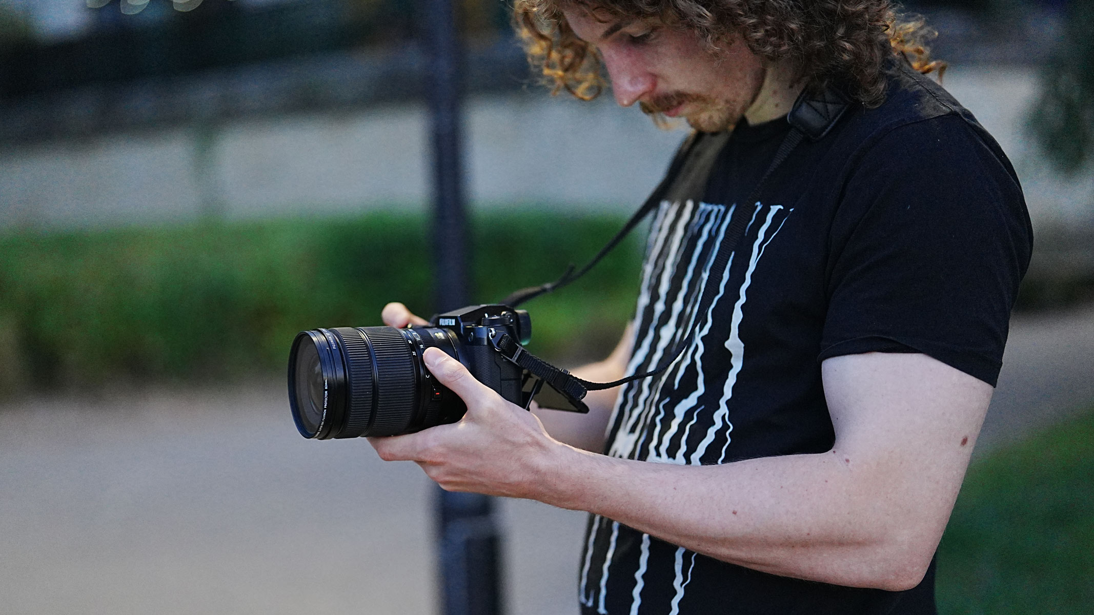 A man holding the Fujifilm GFX100S II whilst looking down at the screen, with bushes in the background.