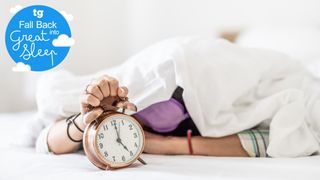 A woman hiding under the duvets while holding an alarm clock after the clock change