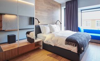 Interior view of a room at Ion City Hotel, Reykjavik, Iceland featuring wood flooring, a wood covered wall, a mirrored wardrobe, a grey bed with white pillows and linen along with a dark coloured fringed throw, windows, a blue window seat and grey curtains