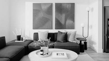 black and white image of living room with white walls, two artwork above modular corner sofa, with cushions, a black side table next to a large orb-style floor lamp, with a white round coffee table on a white rug
