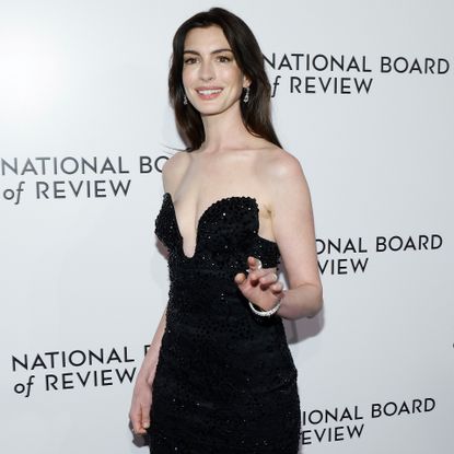 Anne Hathaway is wearing a sleeveless black dress smiling and waving to the camera. 