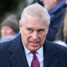 Prince Andrew has white gray hair and wears a large overcoat with a white shirt and red tie underneath