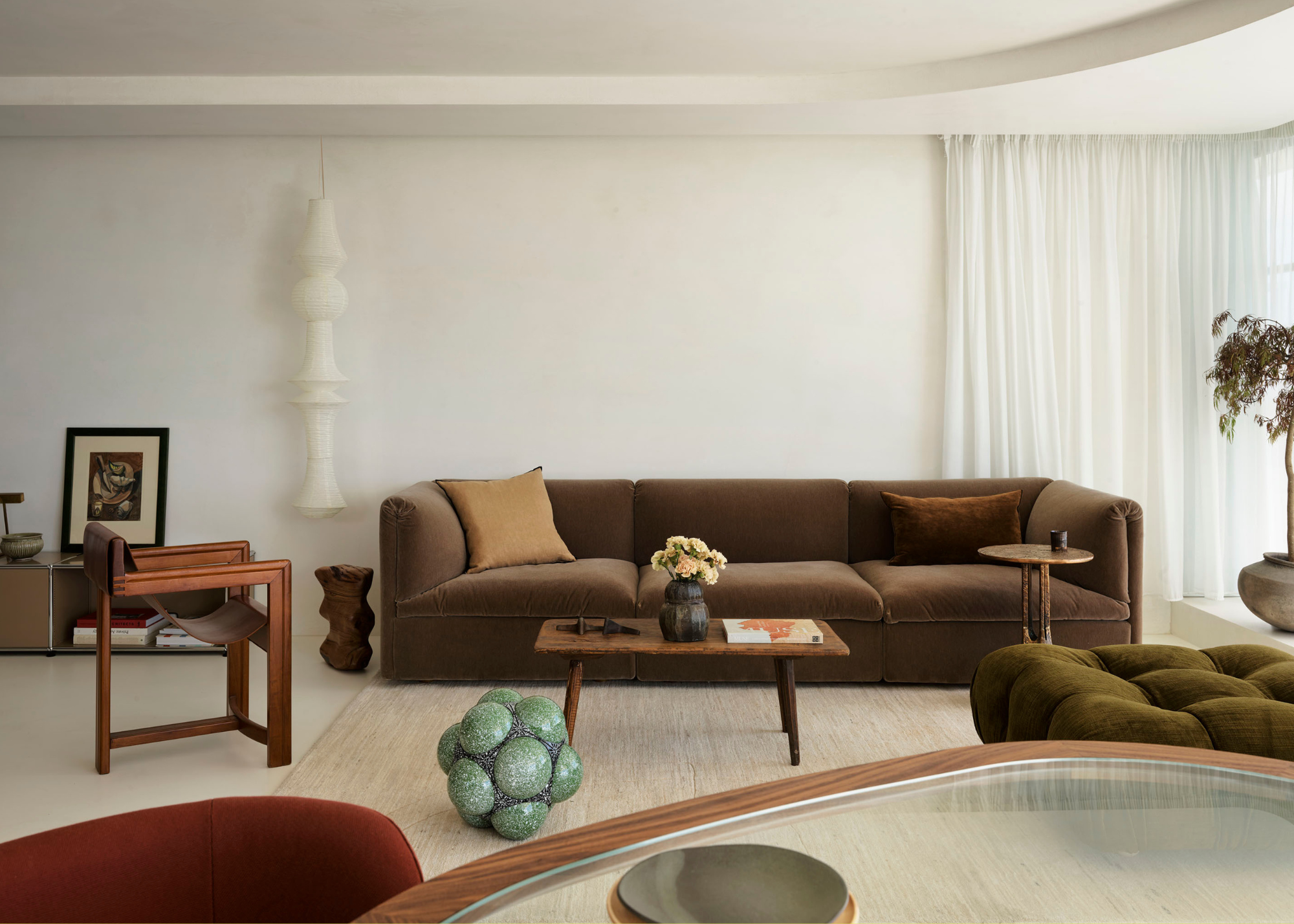 A white living room with a brown couch, a green lounge chair, a white lantern, a wood armchair, a coffee table, and a potted tree