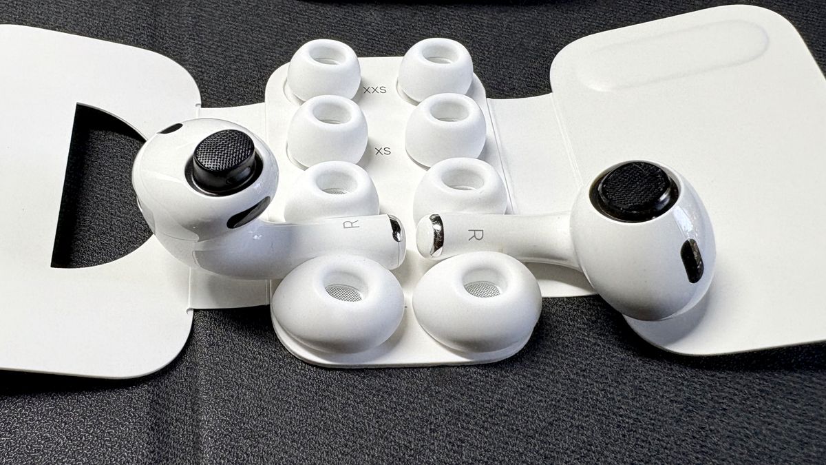 Apple AirPods Pro 3 either worn by a man standing in front of a red-brick wall, or on a table with gray twine cover