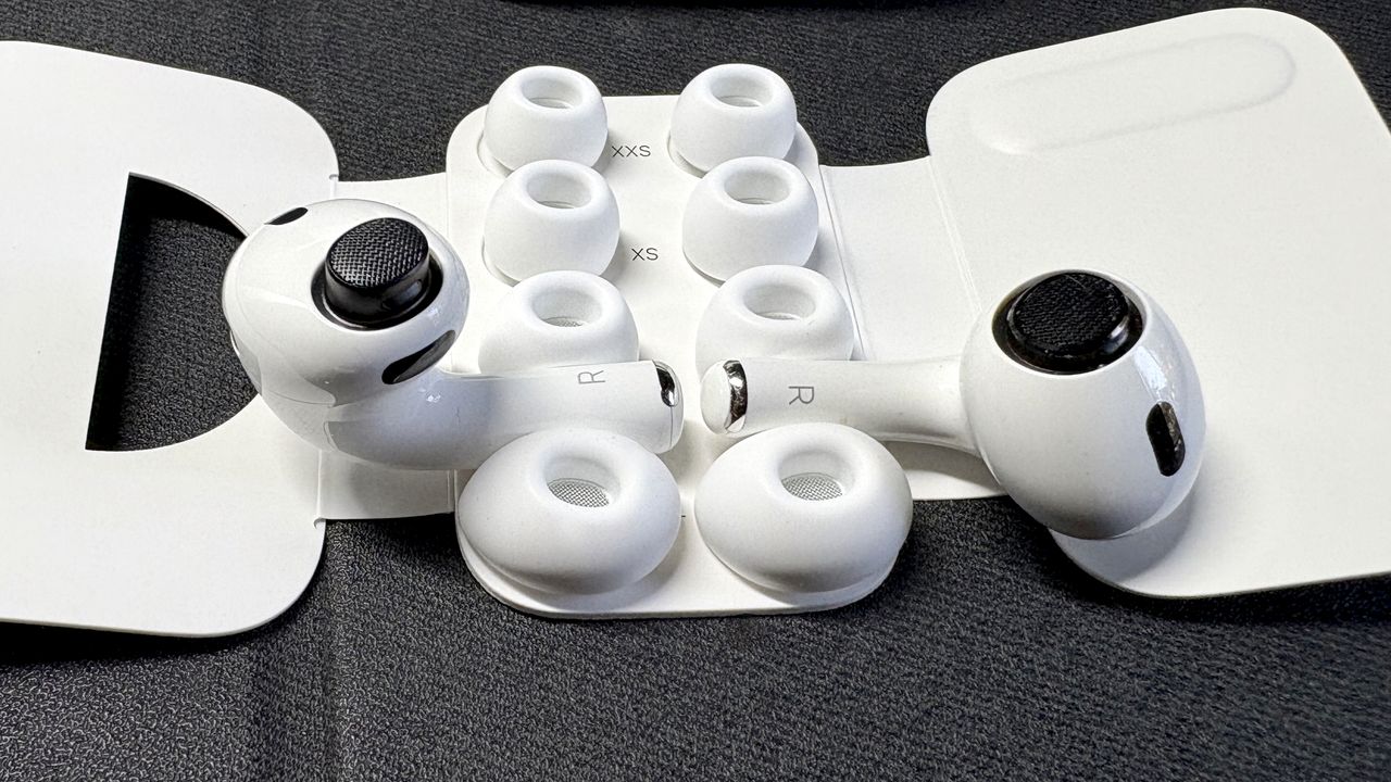 Apple AirPods Pro 3 either worn by a man standing in front of a red-brick wall, or on a table with gray twine cover