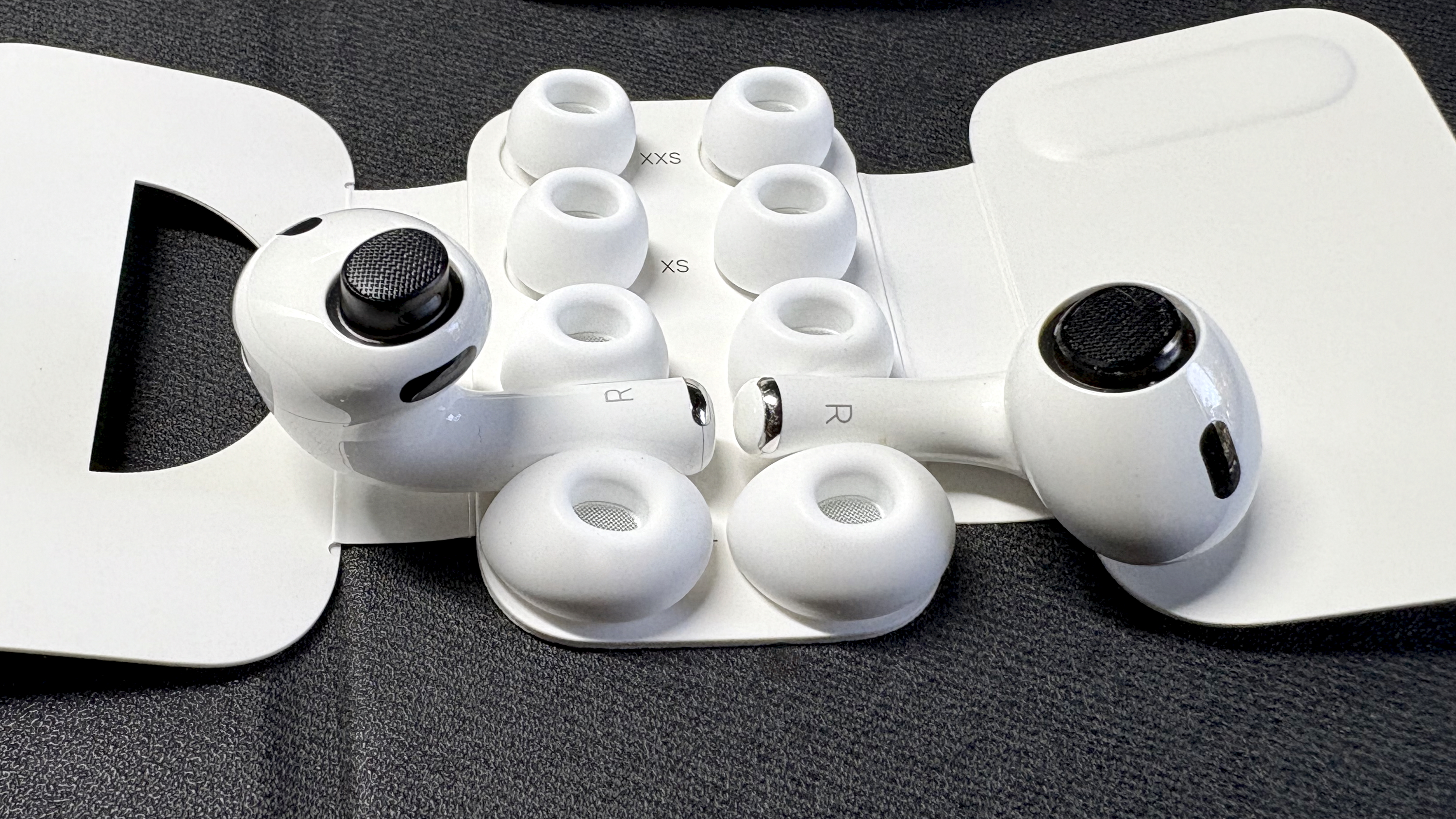 Apple AirPods Pro 3 either worn by a man standing in front of a red-brick wall, or on a table with gray twine cover