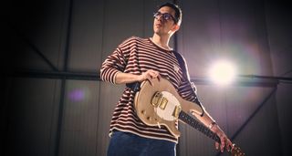 Cory Wong wears a Breton-stripe shirt and poses with his new gold Ernie Ball Music Man StingRay II signature guitar