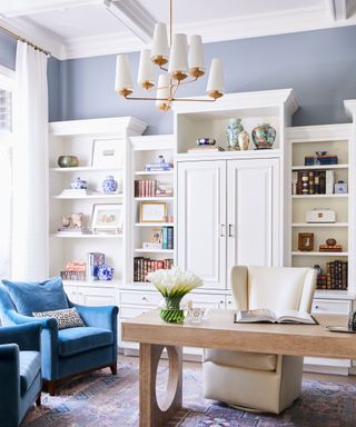 Bright living space with sky blue painted walls and white paneled ceiling, facing large white cabinet with shelving decorated in objects, books and accessories, bras metallic pendant light with white shades, large wooden table with white armchair, two blue armchairs to the left, blue and gray patterned rug