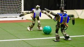 Humanoid robots stand either side of a football in front of a goal post.
