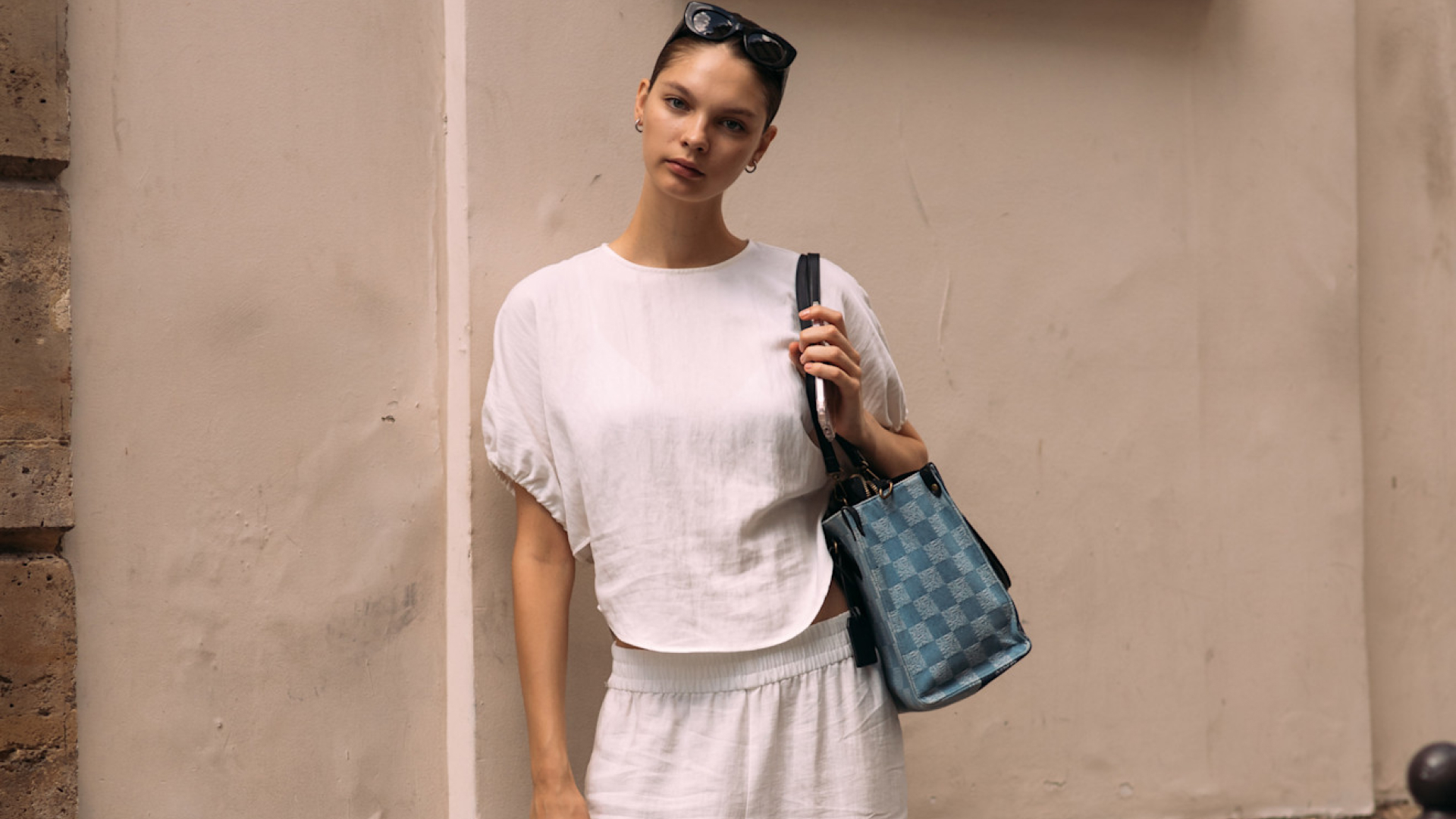 woman wearing white linen set, checkered bag, and sunglasses standing in front of concrete wall