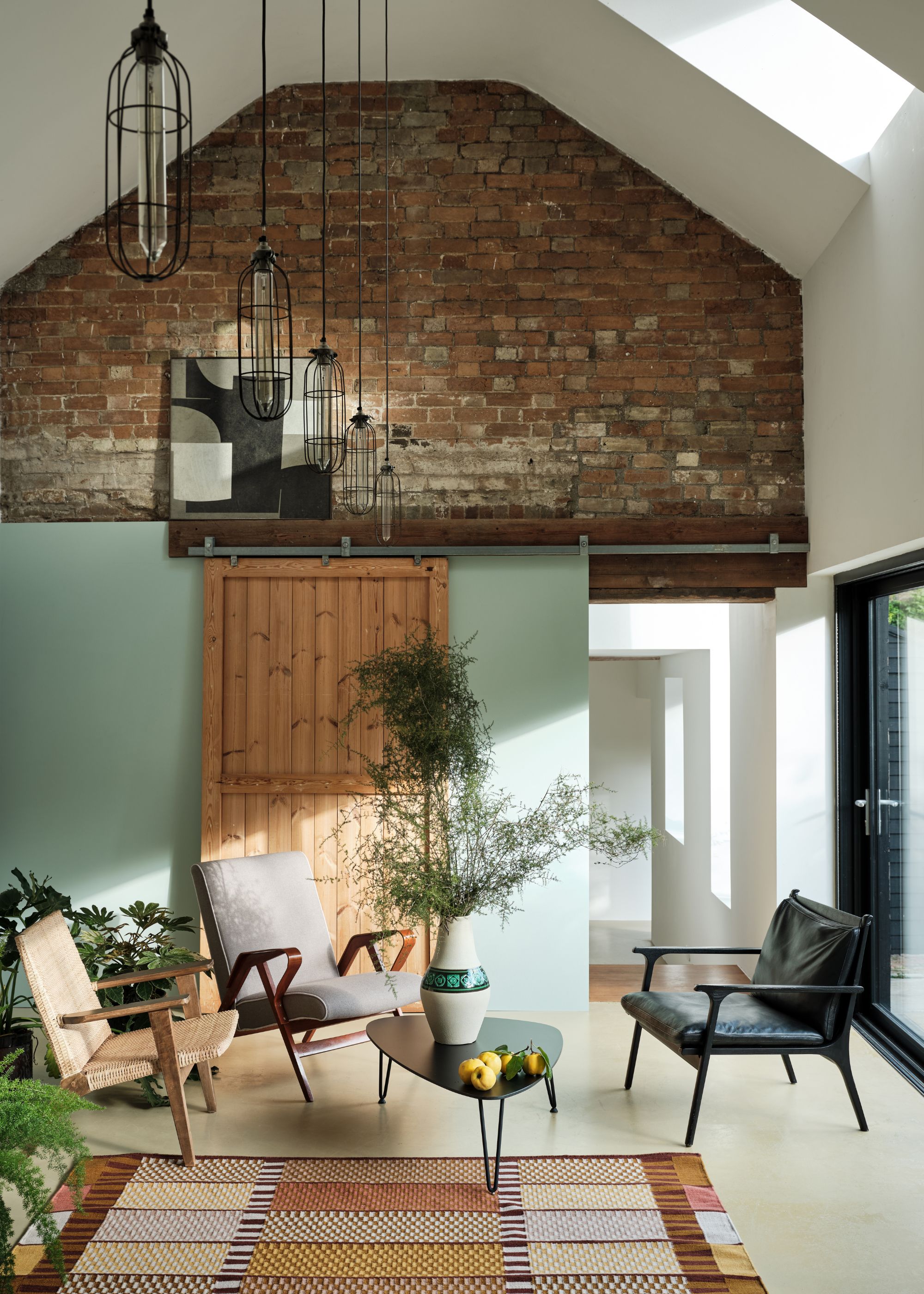 Light and airy living room with sloped high ceilings and exposed brick wall, light green accent wall, and midcentury modern chairs.