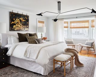 Neutral bedroom with sculptural black pendant light, rug, bench at foot of bed, artwork above bed, seating area beside window