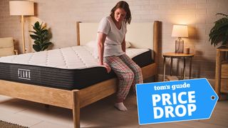 A woman sits on the edge of Plank Firm Mattress on a bed frame in a bedroom, demonstrating the excellent edge support of this mattress. A Tom's Guide price drop deals graphic in the bottom right corner
