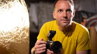 A close-up of a man in a studio holding a Canon camera with a flash radio trigger attached to the hotshoe