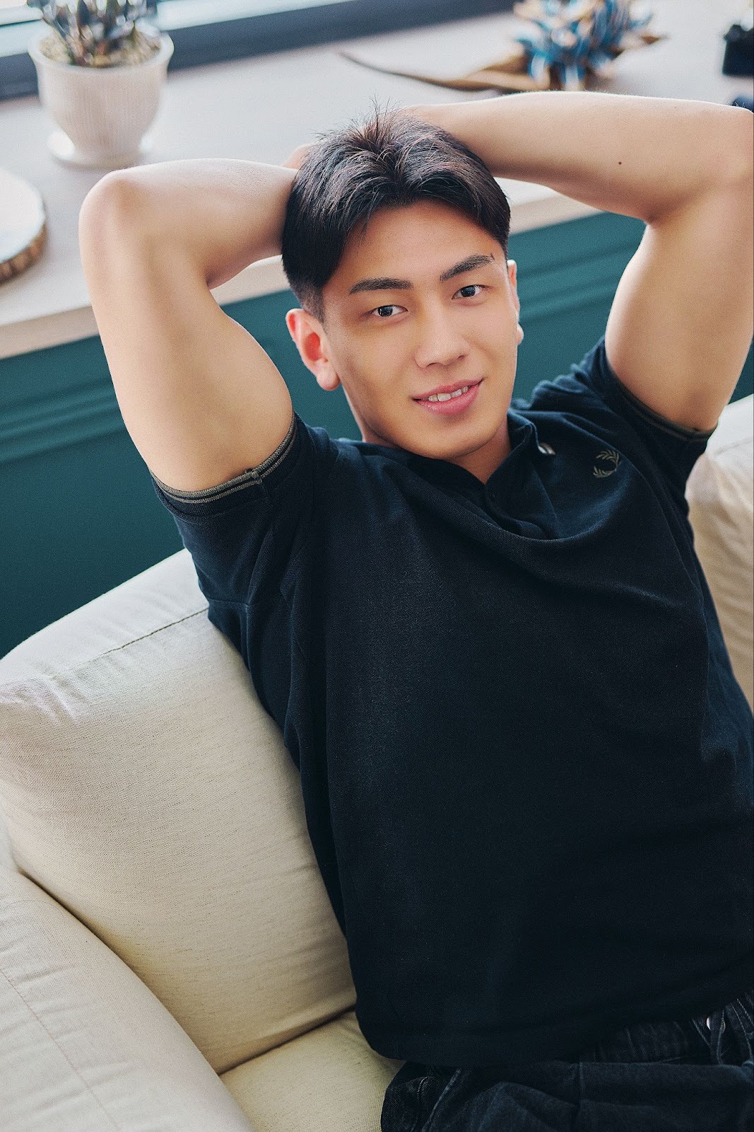 A Thai man named Huwei wears a black t-shirt while sitting on a sofa and posing with his hands behind his head, in a cast portrait for 'The Boyfriend.'