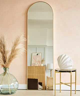 Modern arch design floor length mirror leaning against pale peach wall, surrounded by natural textures and sculptural forms.