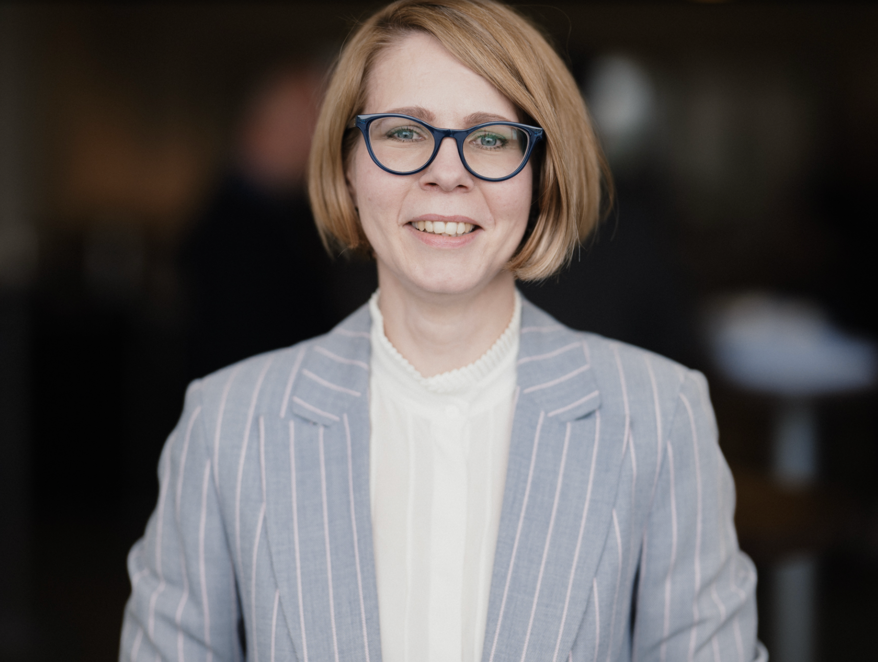 A woman with a blond bob and glasses wearing a gray blazer with stripes.
