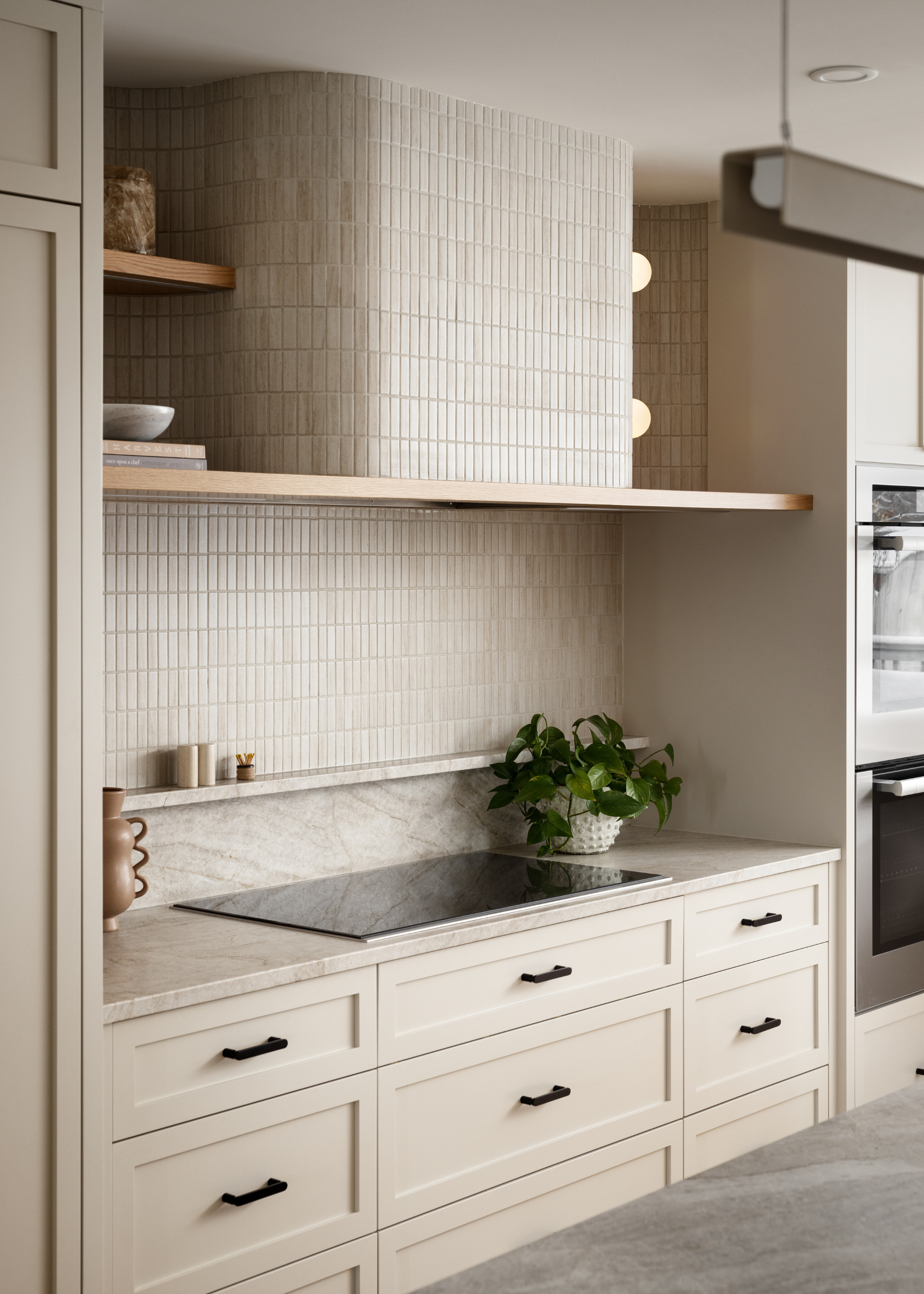 A minimalist kitchen with a tile backsplash, a stone countertop, an induction hob, a potted houseplant, a structured vase, a small ledge, and in-built wooden display shelves