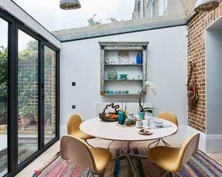 A small kitchen extension with a glass ceiling above round white dining table and retro yellow chairs