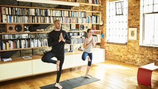 father and daughter do yoga tree pose in loft apartment, in front of a large open shelving unit full of books and records, with large windows to the right