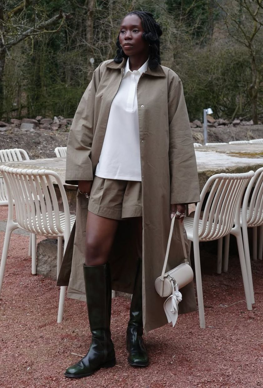 @daniellejinadu wears a long neutral trench coat, white polo sweatshirt, neutral shorts, black riding boots and white barrel bag