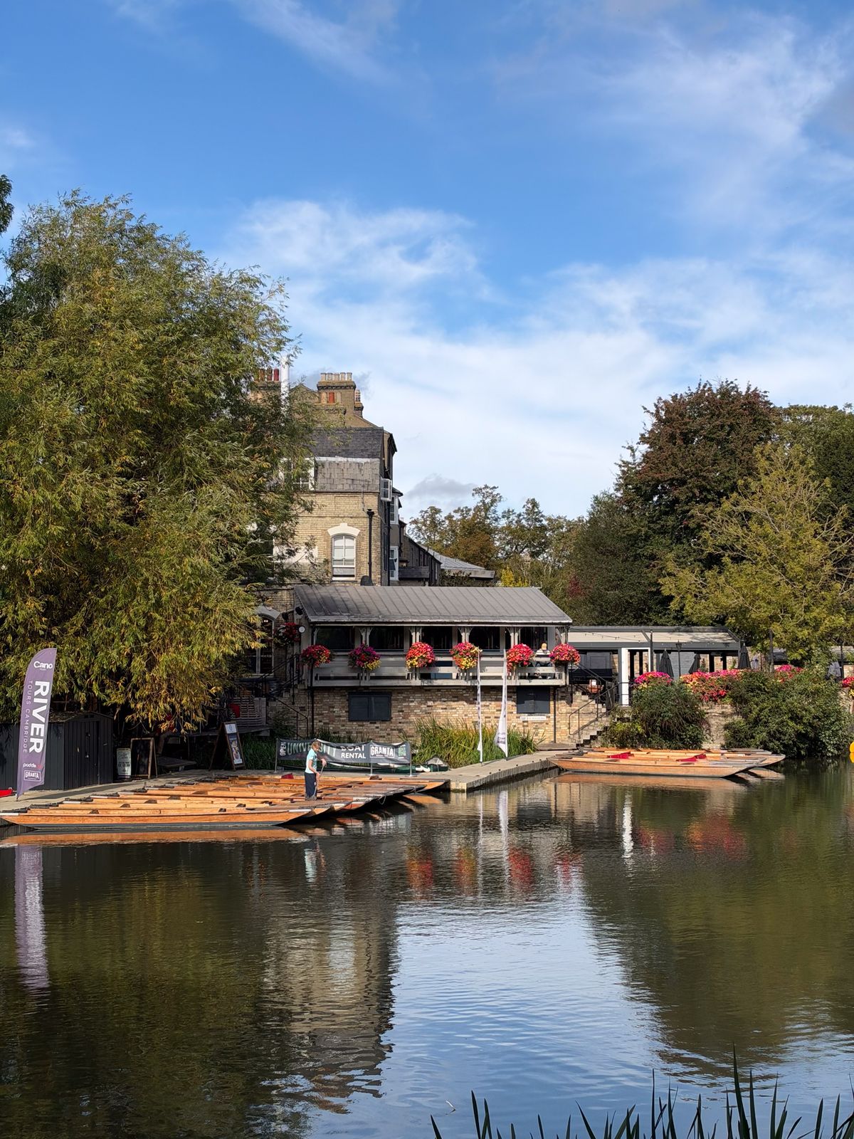 Riverside building with flower-filled balconies and rows of wooden punts reflected in calm water.