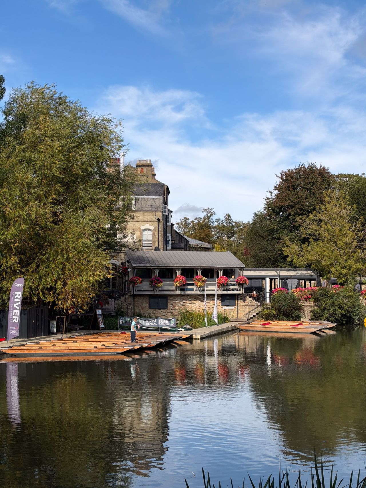 Riverside building with flower-filled balconies and rows of wooden punts reflected in calm water.