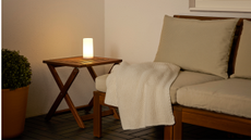 A portable table lamp sits on a wooden side table next to a wooden-based outdoor sofa with beige seat cushions with a white throw draped over the edge in an outdoor seating area at dusk