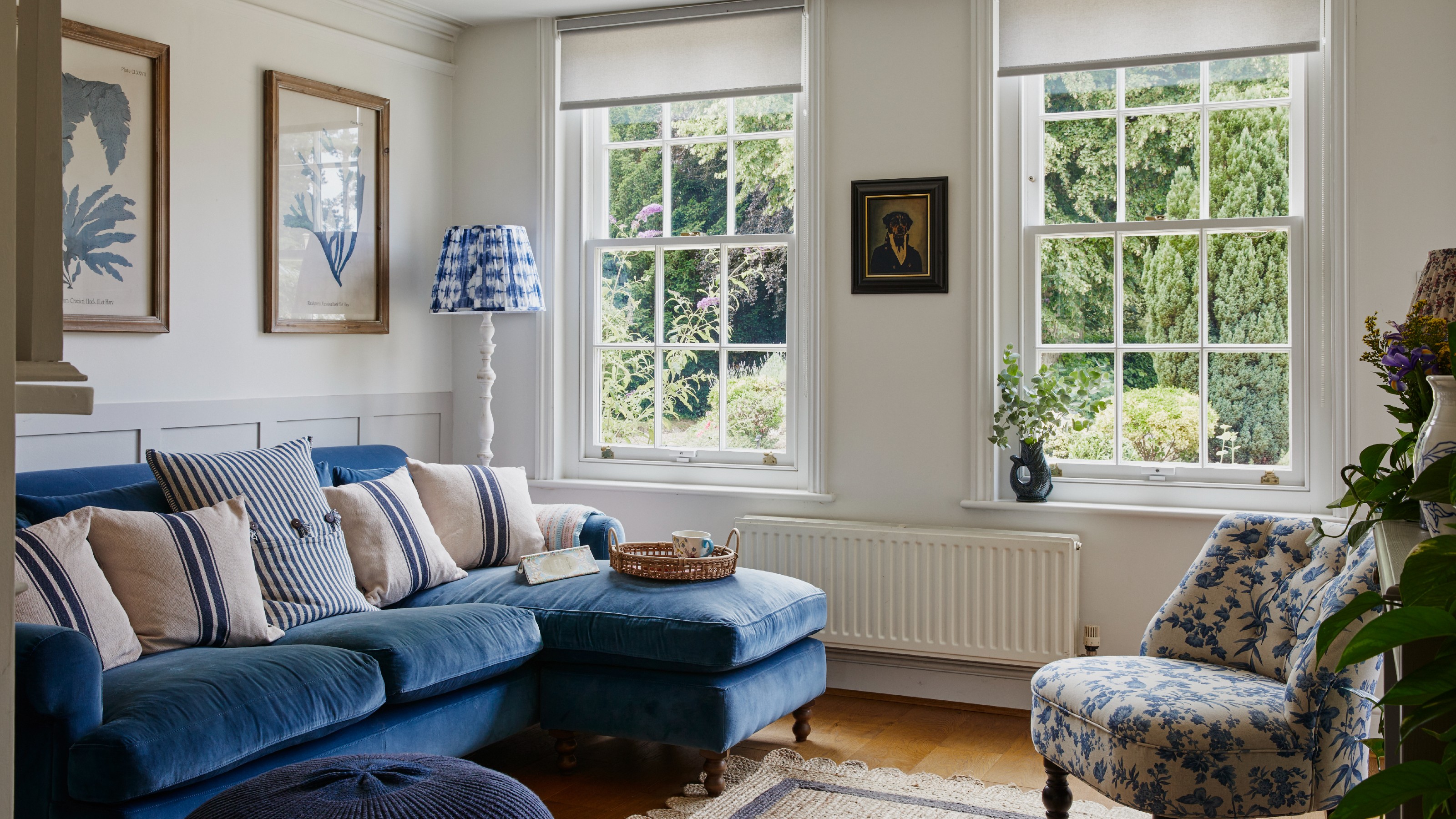 Light-filled living room with artwork on the walls, a blue velvet sofa, blue and white floor lamp, blue and white accent chair, navy footstool and sash windows