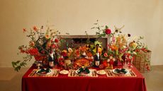 Image of a table set in an extravagant fall theme. There is a red tablecloth, and the center is filled with flower arrangements.