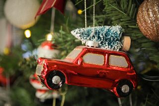 A close up of a red car tree ornament with a small, bottle-brush tree on top, hanging from a Christmas tree branch.