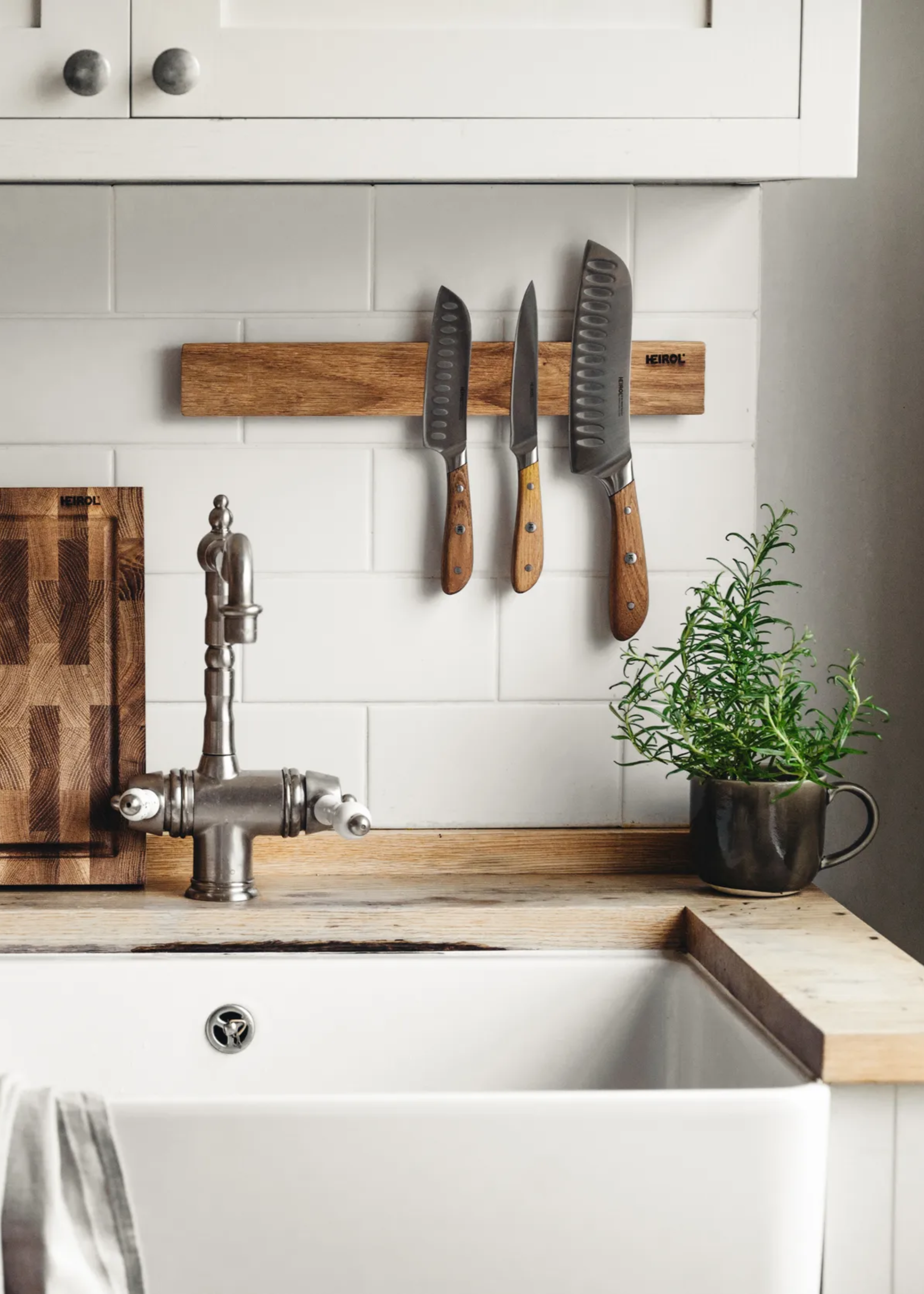 A magnetic knife holder on the wall near the sink, by a chopping board and a rosemary plant in a teacup