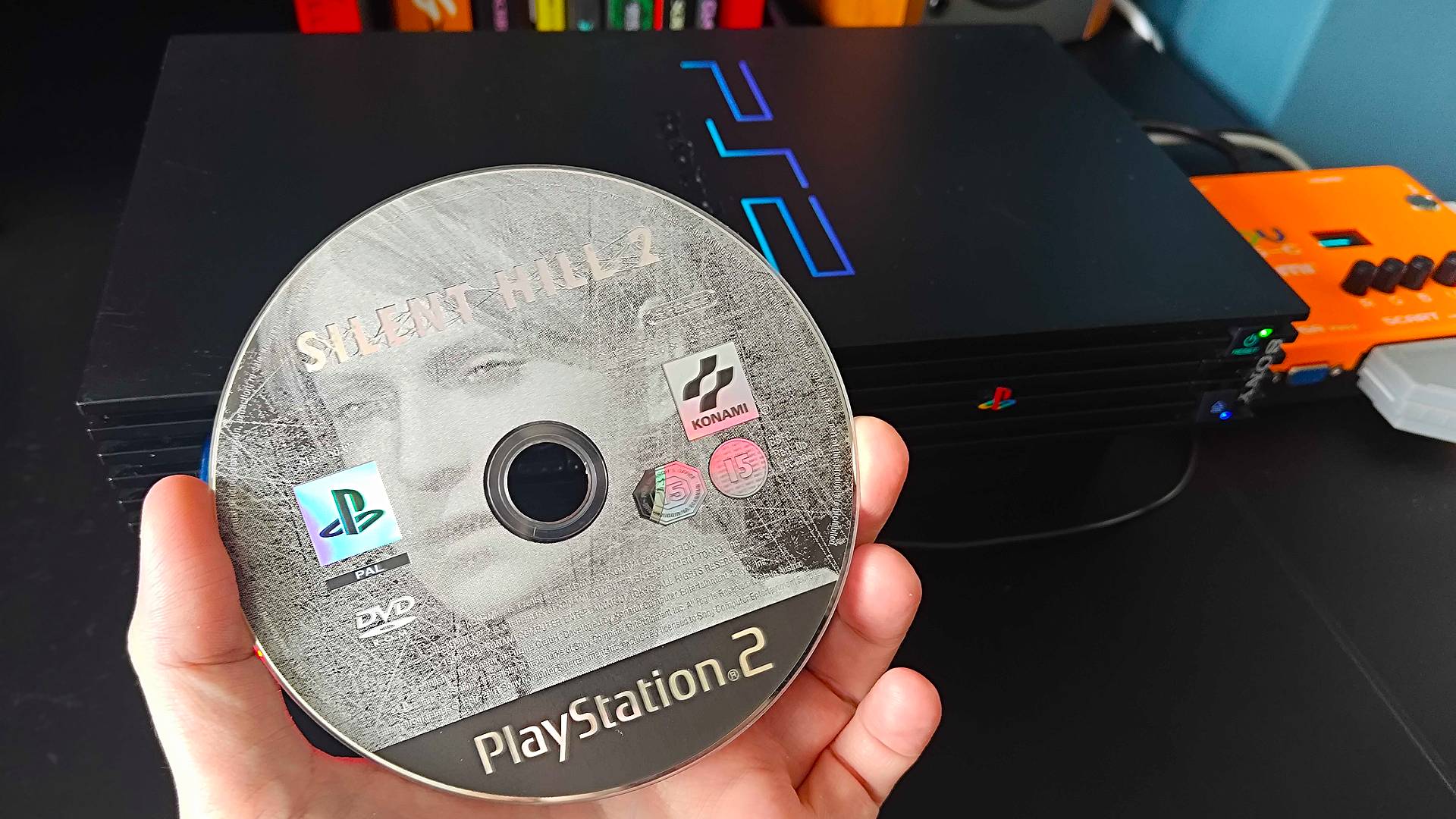 Hand holding Silent Hill 2 disc next to PS2 console.