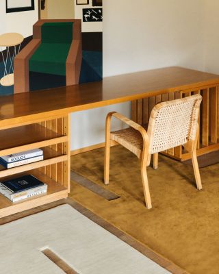 A close-up image of an office space. The desk is natural wood with a minimalist design, and there is a wood and rattan chair in front of it. On the floor, there are two cutout rugs (yellow and white). Above the desk is a contemporary artwork of a chair.