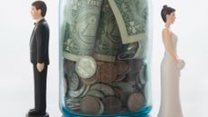 Miniature bride and groom dolls standing on either side of a savings jar full of coins and dollar bills