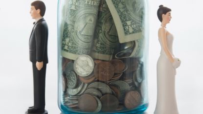 Miniature bride and groom dolls standing on either side of a savings jar full of coins and dollar bills