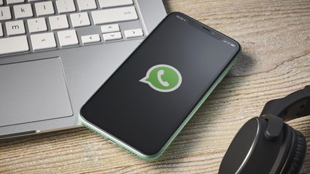 An iPhone with the WhatsApp logo on the screen, sitting on top of a laptop keyboard with a pair of headphones in the corner of the frame, on a wooden surface