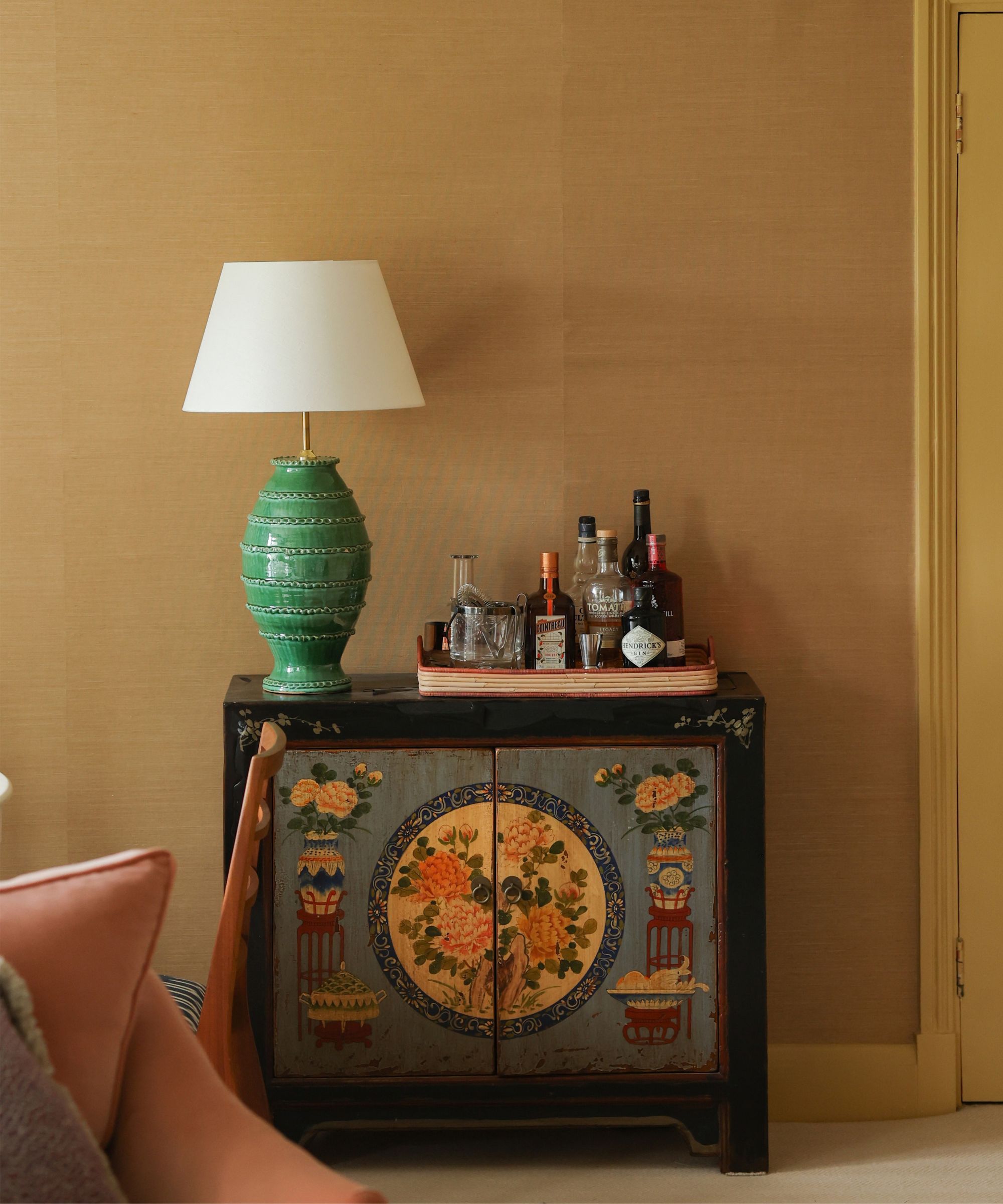 A living room with mustard grasscloth walls, an antique cabinet against the wall, a tray of alcohol and glassware, and a large green ceramic table lamp beside it.