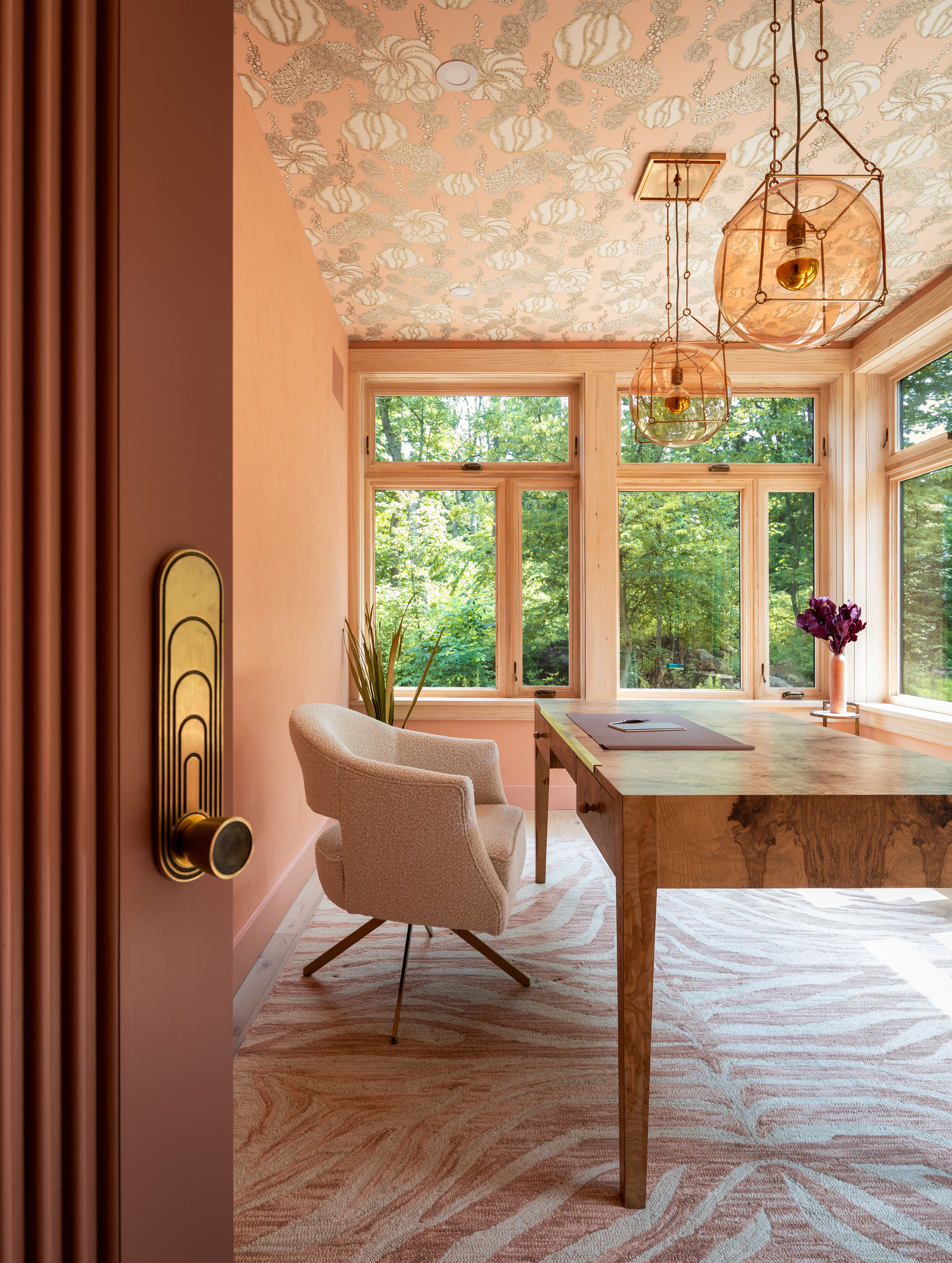 Image of a peach home office with lots of windows and a pink and white zebra rug. The ceiling has patterned wallpaper, and there are two amber glass pendant lights. There is a burlwood desk and a boucle desk chair.