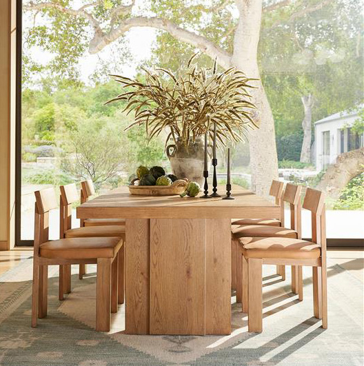 a wooden dining table and chairs next to a full glass window with the sun shining in with a large vase of foliage on the table