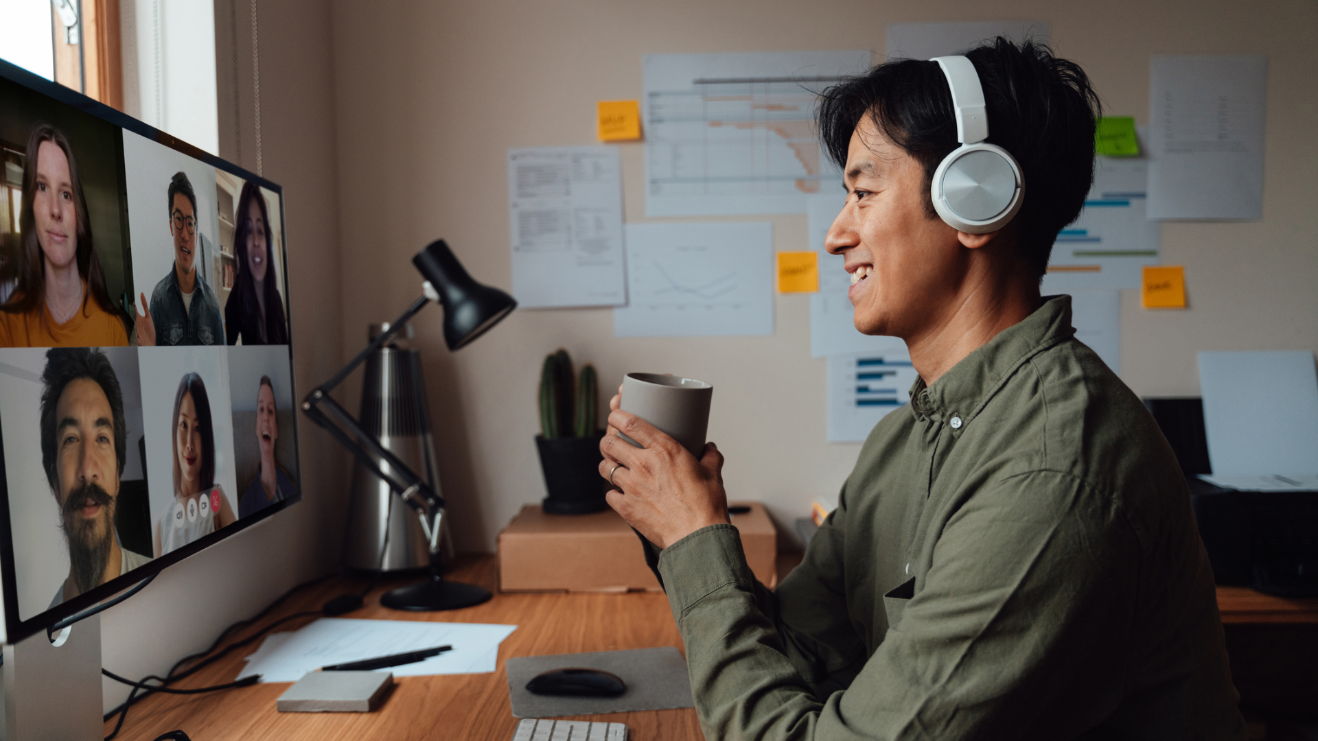 A man holding a coffee mug at a desk in a home office conducting a video call with six team members