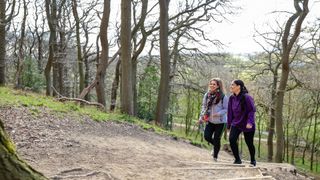 Two women wearing activewear and hiking backpacks, laughing while walking up a muddy path in the forest
