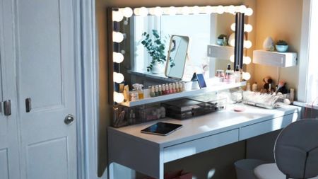 A fully built-out vanity setup with a large Hollywood-style mirror with bulb lighting, white desk drawers and makeup and skincare products