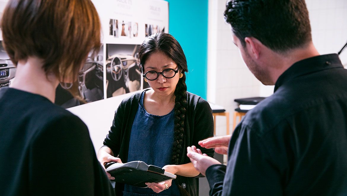 Image of two women and a man stood having a conversation, front facing women holding an item of discussion, white and blue walls, images of car interiors on the left wall 