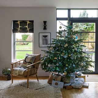 Wrapped presents beneath Christmas tree decorated with ornaments and blue fairy lights next to chair in living room