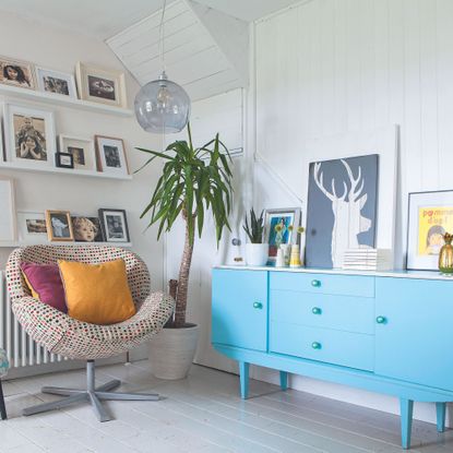 Hallway with painted floorboards, blue sideboard, white shelves displaying framed photographs, armchair and footstool. Gill and Sandro Deriu's renovated three bedroom detached cottage in Cork, Ireland.