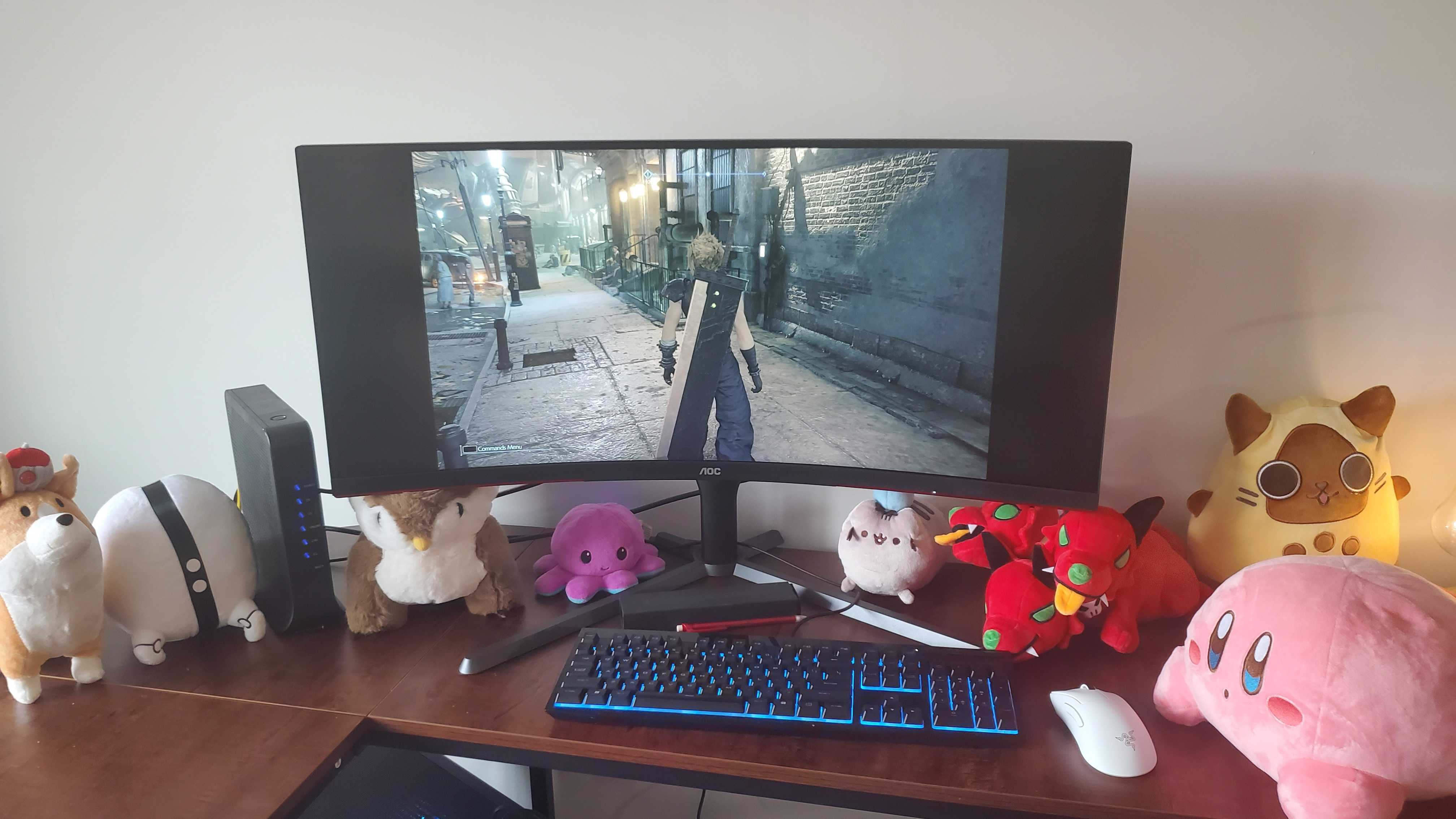 a large curved monitor on a wooden table with various plushies under it