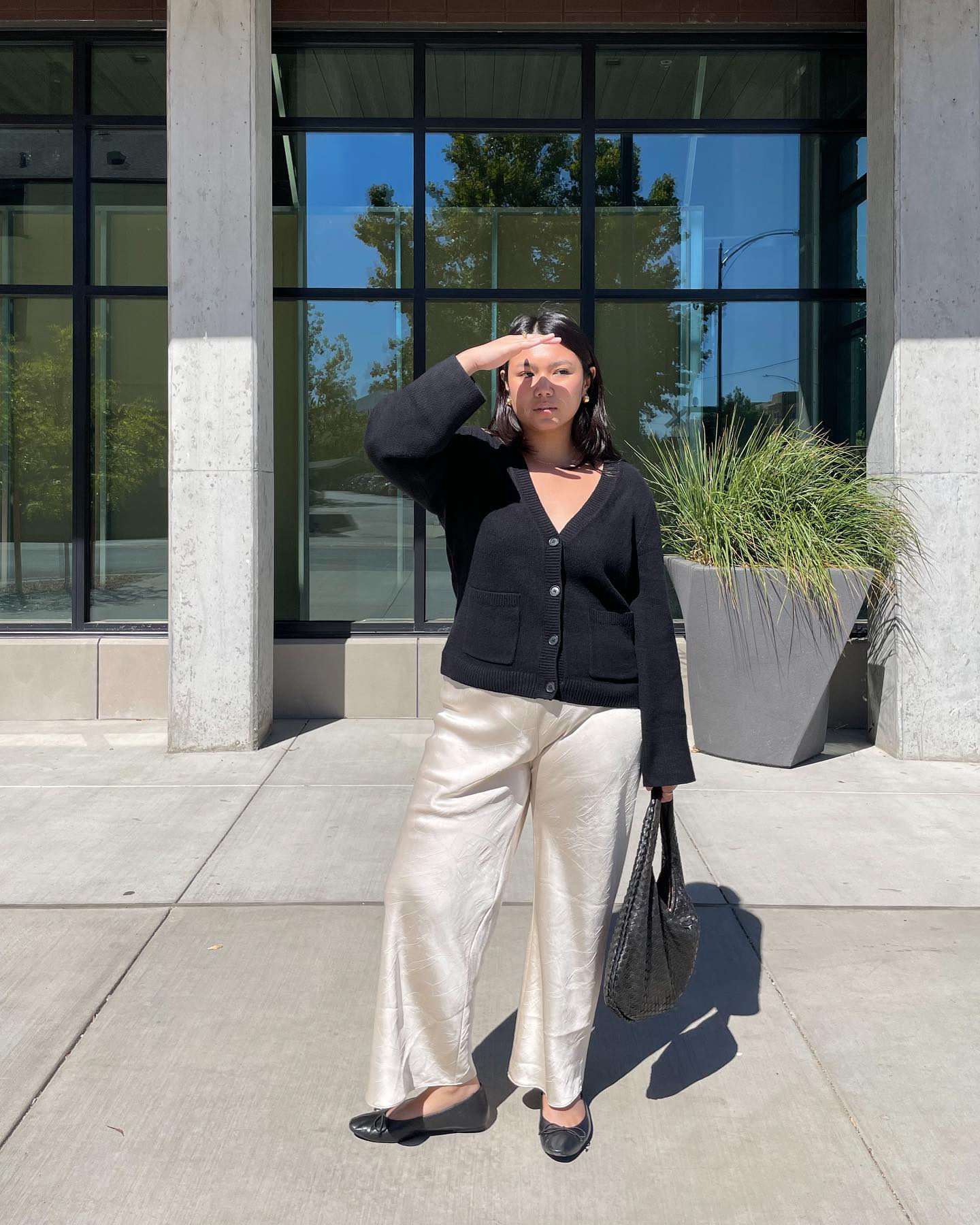 An image of an influencer wearing a black button-down cardigan with cream-coloured satin trousers and black ballet flats carrying a black woven tote