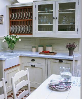 A glass kitchen cabinet displaying glassware in a traditional white kitchen with plate racks and a wooden table