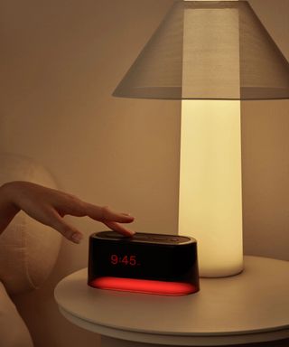 A hand pressing the snooze button on a Loftie alarm clock; in the background, a lamp on a nightstand against a brown wall.